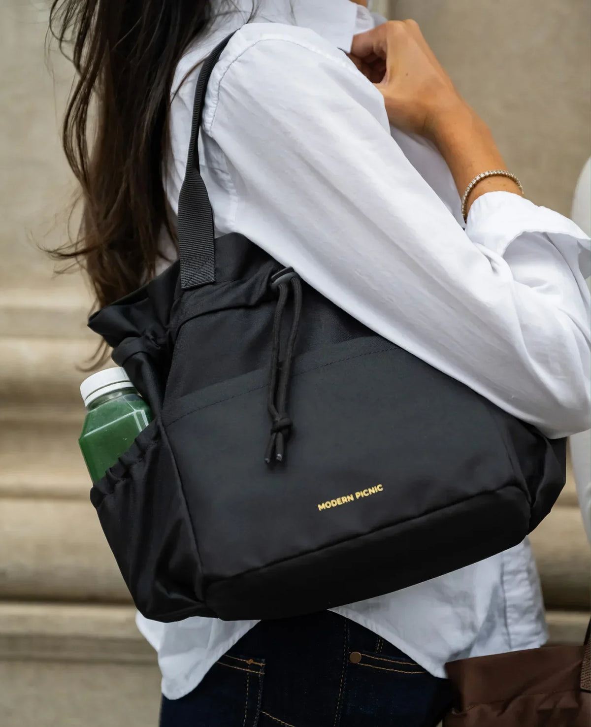 Woman wearing a white shirt carrying a black Modern Picnic insulated tote bag with a green drink.