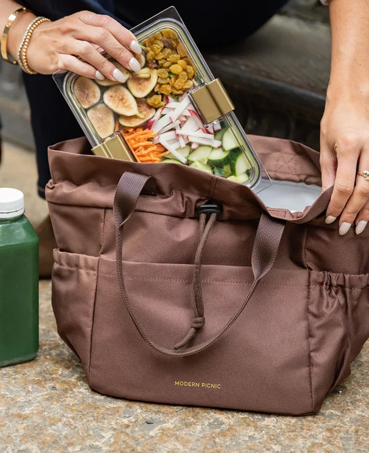 A person placing a clear container of healthy snacks into a brown Modern Picnic insulated tote bag.
