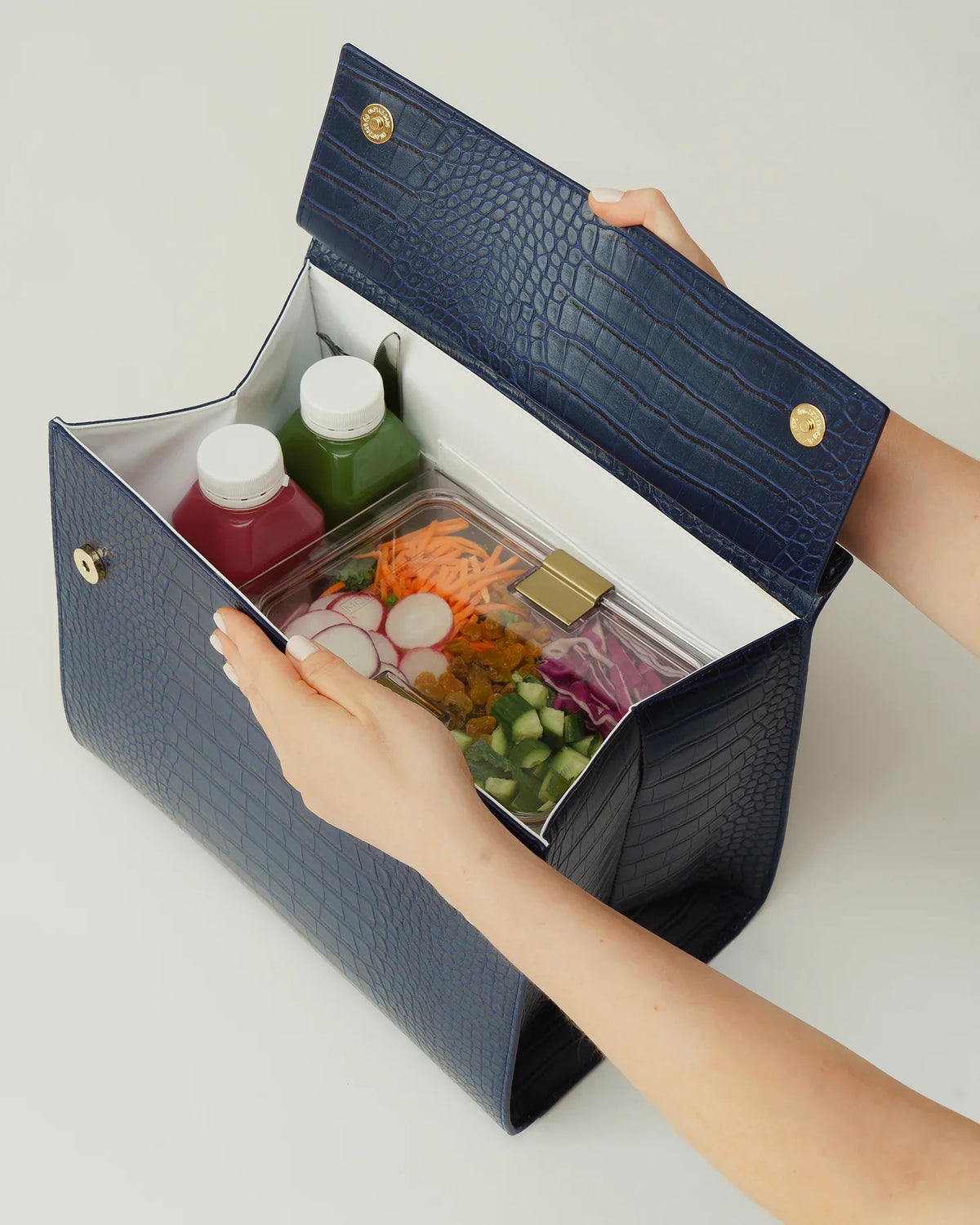 Hands opening a navy blue croc-embossed insulated lunch tote containing salad and juices.