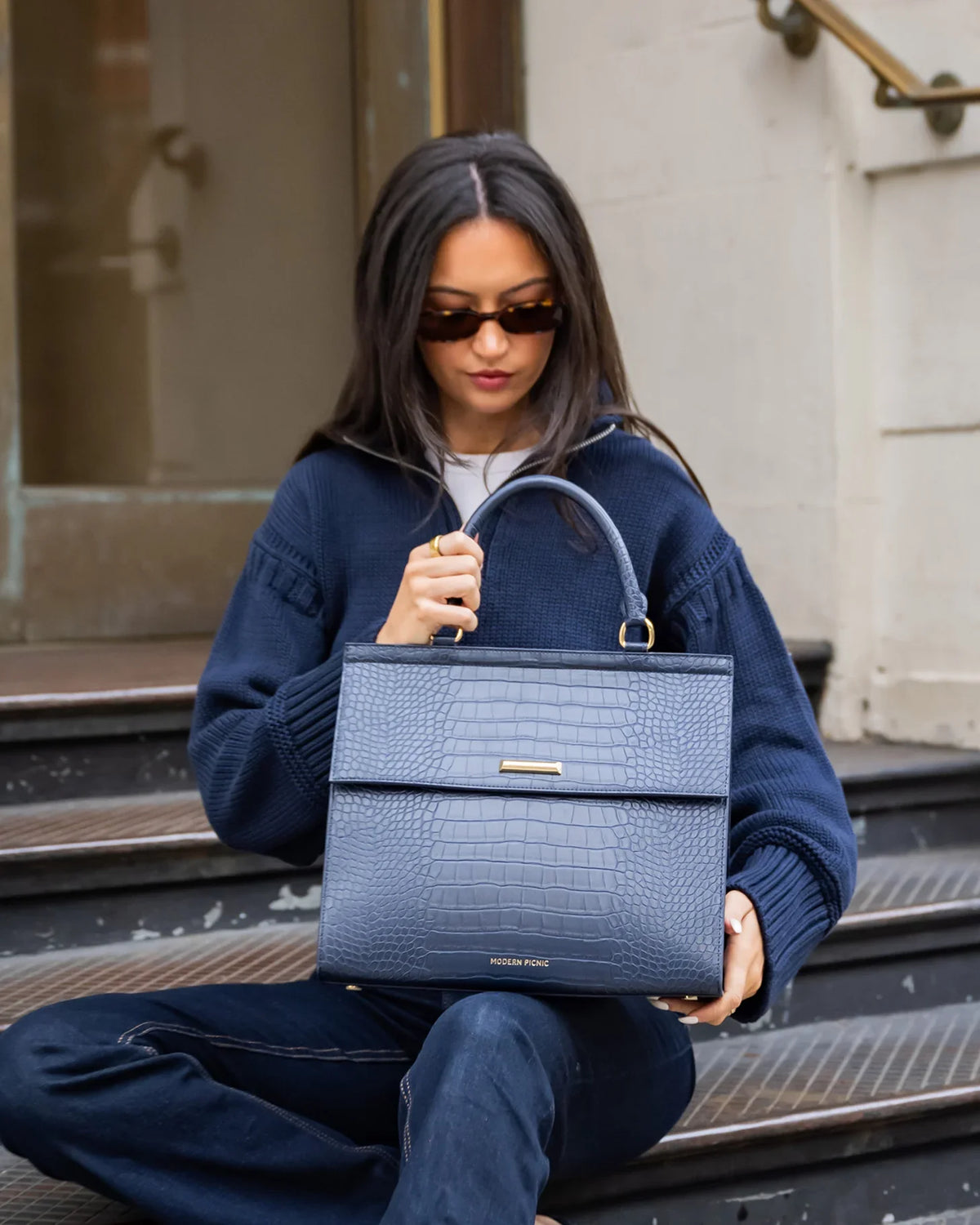 Woman sitting on stairs holding a navy blue crocodile-textured vegan leather lunch bag.