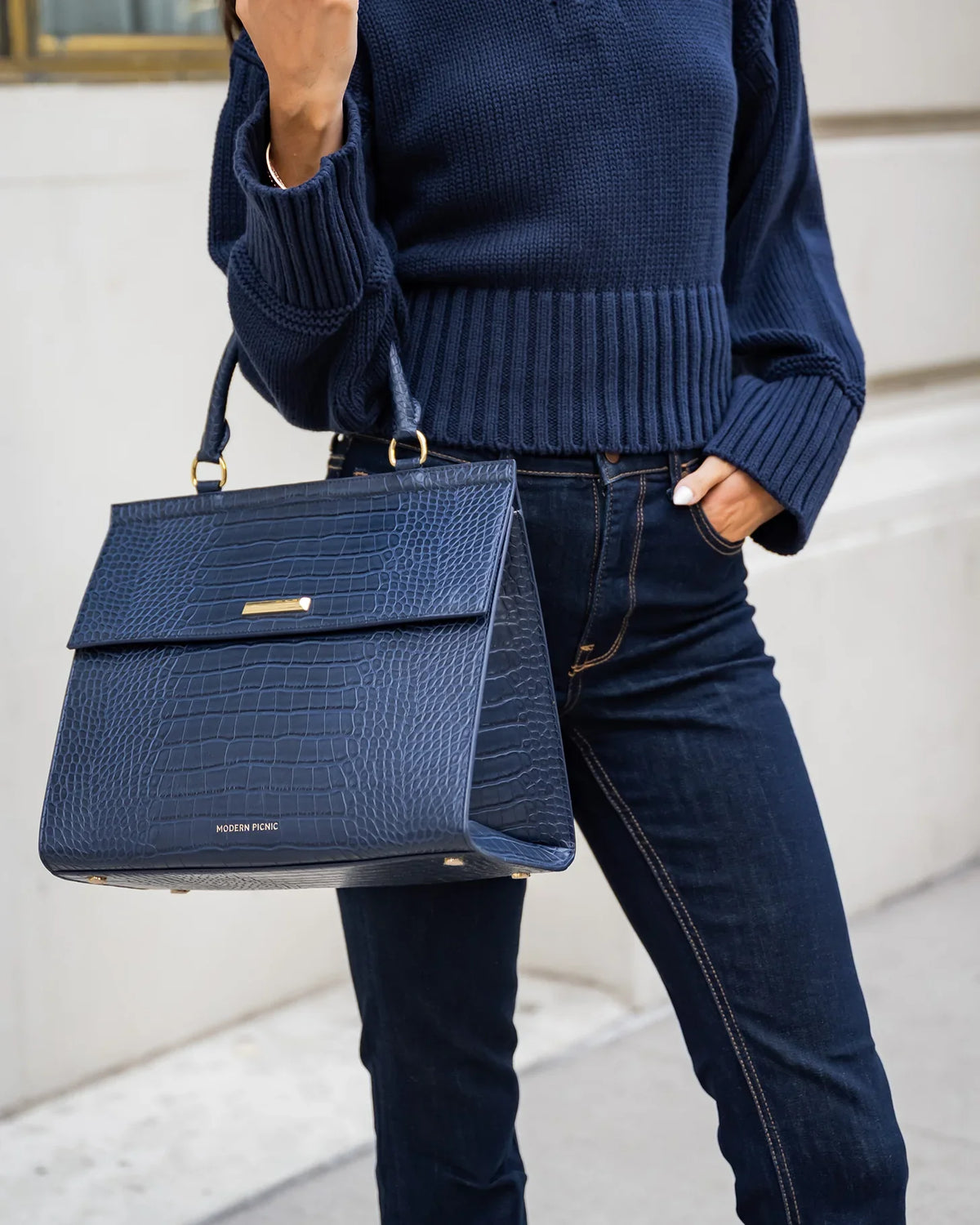 Woman wearing a navy sweater and jeans holding a navy blue croc-embossed insulated tote bag.