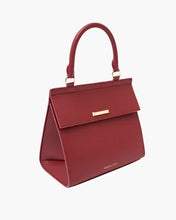 A structured red smooth luncher with a curved top handle is shown at a side angle, featuring a flap closure detailed with a small horizontal gold-toned bar and matching gold hardware on the handle attachments, all set against a clean white background.