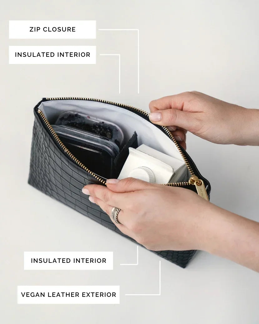 A close-up, overhead view of the black pouch with the zipper open. Hands are opening the bag, revealing the white insulated interior packed with a small juice box and a clear container of dark berries. Labels point out the zip closure, insulated interior, and vegan leather exterior.