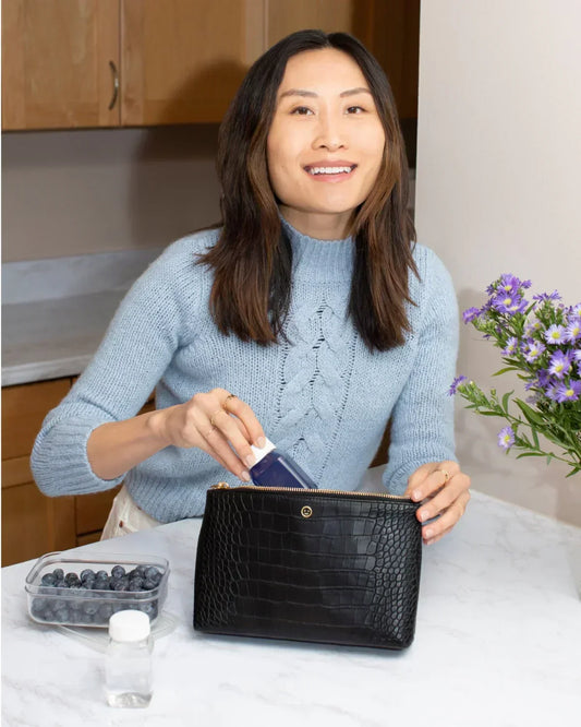 A smiling woman in a light blue cable-knit sweater is standing at a marble kitchen counter, placing a small dark bottle into an open black pouch with a crocodile-embossed texture. A clear container of blueberries is visible next to the bag.