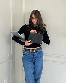 A young woman wearing a black long-sleeve top and blue jeans with a black belt stands against a white wall, holding a laptop in one arm and tucking a black pouch into the other. The pouch has a subtle crocodile-embossed texture.
