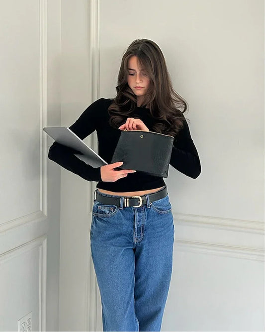 A young woman wearing a black long-sleeve top and blue jeans with a black belt stands against a white wall, holding a laptop in one arm and tucking a black pouch into the other. The pouch has a subtle crocodile-embossed texture.