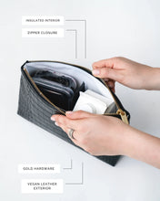 A close-up, overhead view of the black pouch with the zipper open. Hands are holding the bag, revealing the white insulated interior packed with a white juice box and a container of dark berries. Labels point out the insulated interior, zipper closure, gold hardware, and vegan leather exterior.