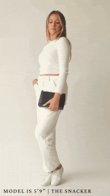 A full-body animation shows a blonde woman wearing a white long-sleeve top and white trousers modeling the small, rectangular pouch. She holds the black pouch by her side, then the animation shows her opening the zipper to reveal the interior.