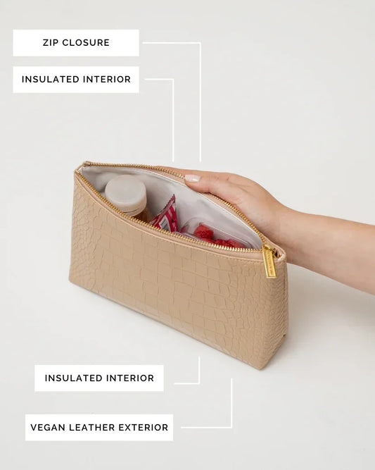 A close-up, overhead view of the cream pouch with the zipper open. A hand is holding back the flap, revealing the white insulated interior packed with a small bottle and fresh raspberries. Labels point out the zip closure, insulated interior, and vegan leather exterior.