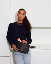 A woman with a long, curly ponytail, wearing a navy sweater and light-wash jeans, leans against white kitchen cabinets. She holds a small black pouch with an embossed texture and a zipper, with a snack bar sticking out.