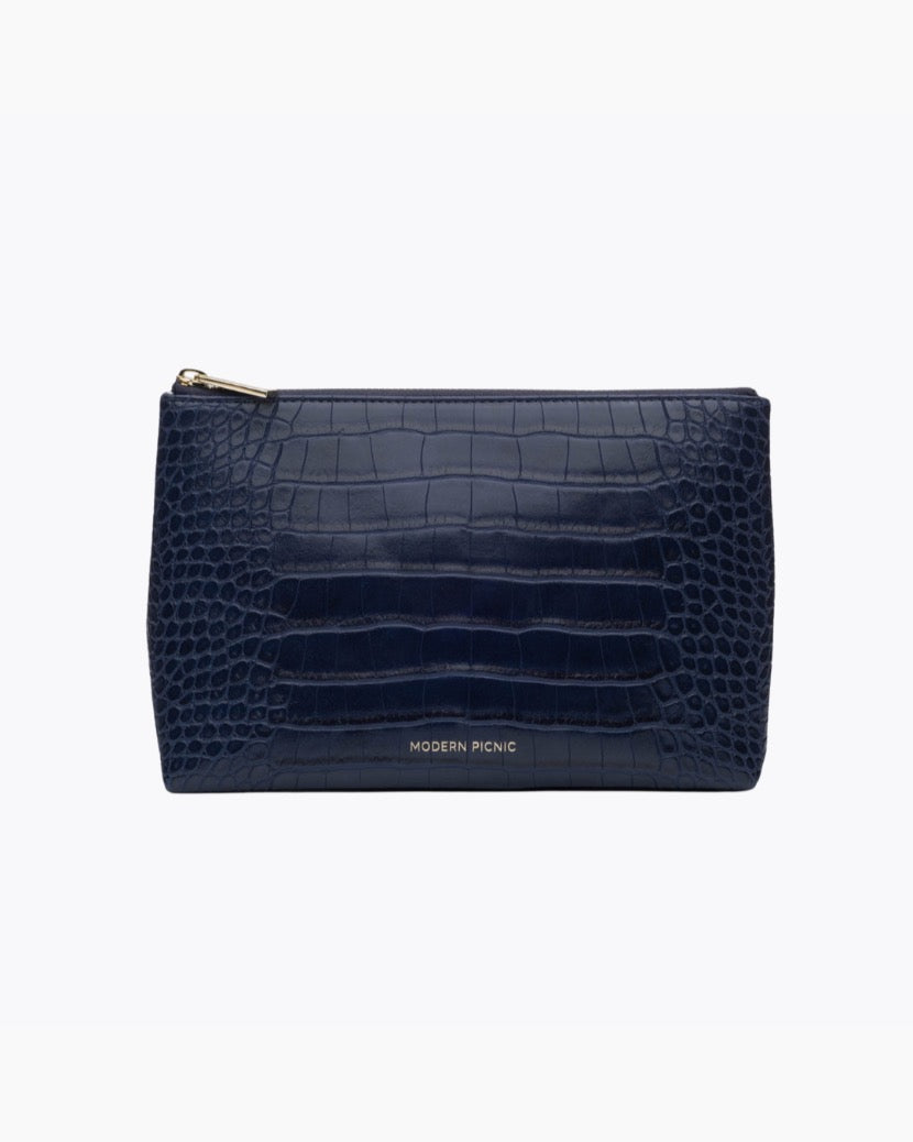 A front-facing studio shot of a small, rectangular navy blue pouch. The bag is made of crocodile-embossed vegan leather and features a gold zipper along the top. The gold 