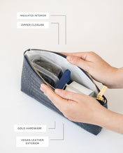 A close-up, overhead view of the navy blue pouch with the zipper open. Hands are opening the bag, revealing the white insulated interior packed with a clear container, blue snack bar, and a white carton. Labels point out the insulated interior, zipper closure, gold hardware, and vegan leather exterior.