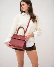 A woman wearing a white sweater and brown shorts holds the structured red smooth luncher by its top handle, with the long adjustable strap draped over her shoulder, demonstrating the bag's size and style against a white background.