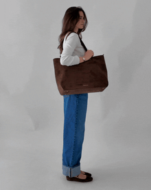 Short video clip of a female model wearing a white long-sleeve shirt and wide-leg denim jeans, confidently posing and walking while carrying a dark brown suede tote bag.
