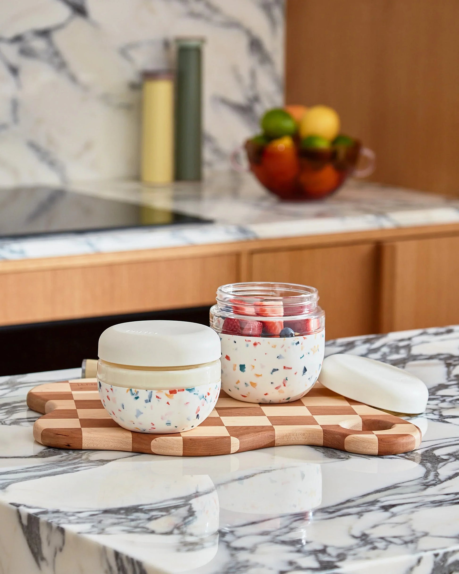Two cream terrazzo food storage bowls with lids on a checkered cutting board in a modern kitchen