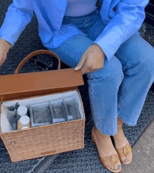 An animation shows a person wearing a blue shirt and jeans sitting down and opening a structured brown woven wicker handbag with a brown vegan leather flap. The interior reveals the white insulated lining and clear containers packed with food and juice bottles.