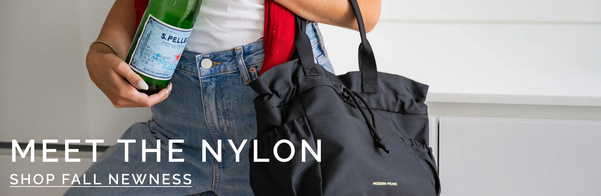 Person holding a black nylon tote bag and a green glass bottle.