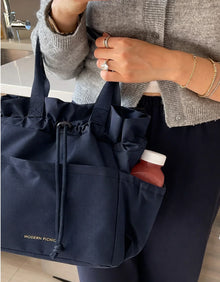 Navy blue insulated lunch tote with drawstring closure, holding a bottle, styled for work or travel.