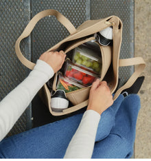 Modern Picnic beige tote bag filled with lunch containers, water bottle, and essentials beside a person sitting on steps.