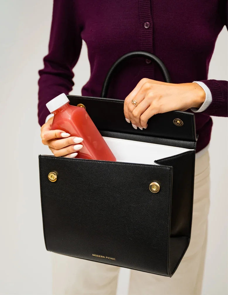 A person holding a black vegan leather insulated lunch bag with a red juice bottle inside.
