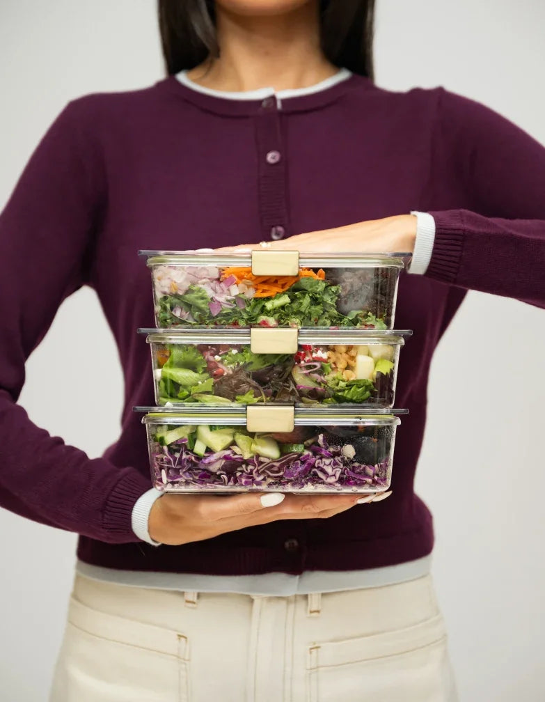 Woman holding three stacked clear meal prep containers filled with fresh salads.