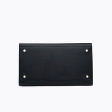 A close-up view of the bottom of the large black handbag, showing the flat, structured base made of pebbled vegan leather. The base is protected by four small gold metal feet (studs), one in each corner.