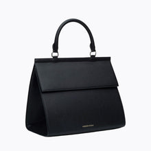 A three-quarter studio shot of a structured large black handbag made of pebbled vegan leather. The bag features a curved top handle, a prominent flap closure, and a visible gold "MODERN PICNIC" logo near the bottom front edge.