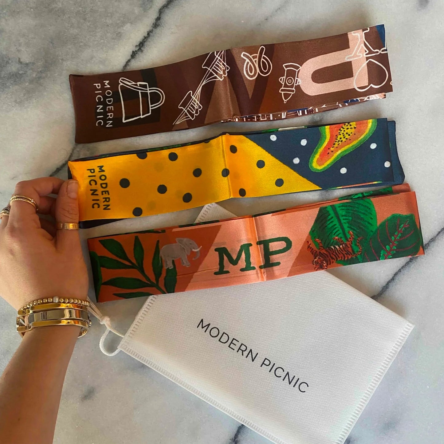 Three Modern Picnic printed bag scarves displayed on a marble surface, featuring the brown-colored scarf, the yellow fruit print scarf, and the tropical MP monogram scarf, with a hand holding one scarf and a Modern Picnic white pouch placed underneath.